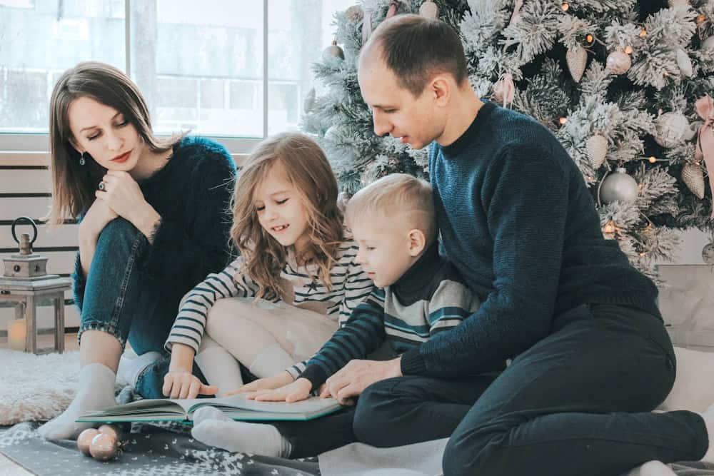 girl and boy reading book sitting between man and woman beside Christmas tree. holiday homeschooling